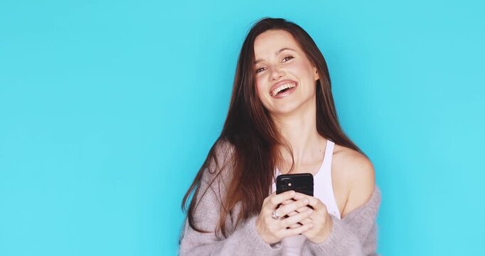 Young woman looking at smartphone with a gentle smile and then laughing, happy facial expression, holding mobile device, engaging with digital content, portrait isolated on blue background, tech use