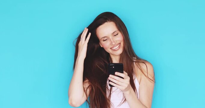 Young brunette woman looking at smartphone with joyful smile and happy expression, laughing while browsing mobile phone, studio isolated on blue background, concept of success and positive connection