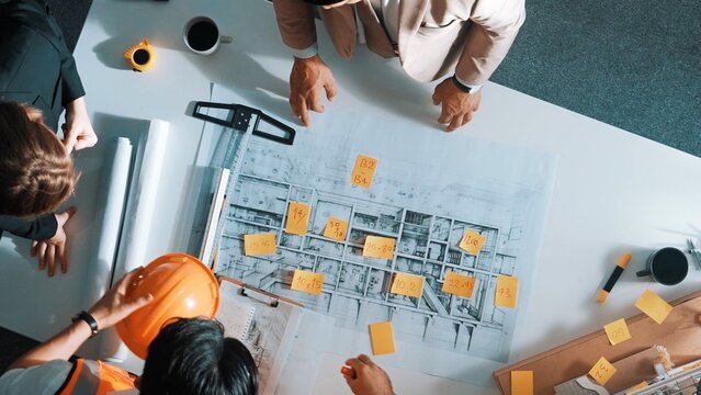 Top down view of architect engineer writing building structure idea on note and stick on blueprint. Aerial view of manager working on notes while consulting team about house structure. Alimentation.