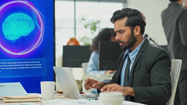 Indian male marketer using laptop and AI consultant on multiple screens to restructure business and marketing strategy in modern open office, South Asian professional digital transformation concept
