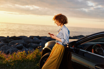 Fototapeta premium Young woman with curly hair stands beside a dark car at sunset by the coast, looking at her phone with a calm, focused expression.