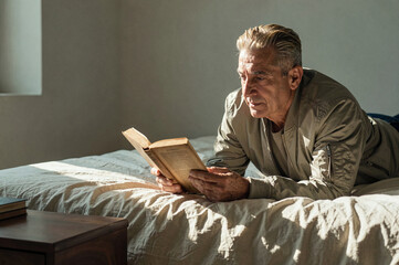 Fototapeta premium Man reads a paper book while lying on an unmade bed in soft side light from a nearby window. His grey bomber jacket contrasts with the wrinkled white linen bedding and neutral wall.