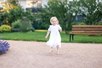 Fototapeta premium Beautiful caucasian 2-year-old toddler girl in a white summer dress walking in a lush green garden in Kladno, Czech Republic, enjoying a sunny June day. Sweet candid moment of childhood, innocence