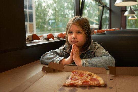 A portrait of a young school-age girl sitting at a table in a pizzeria. A slice of pizza is on the table in front of her.