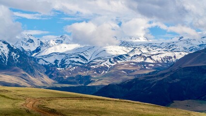 Fototapeta premium Dzhily Su, Russia - June 4, 2026: Scenic view of snow-covered peaks in the Caucasus Mountains near Elbrus Mountain, with rugged mountain ridges, cloudy weather, highland landscape, and panoramic views