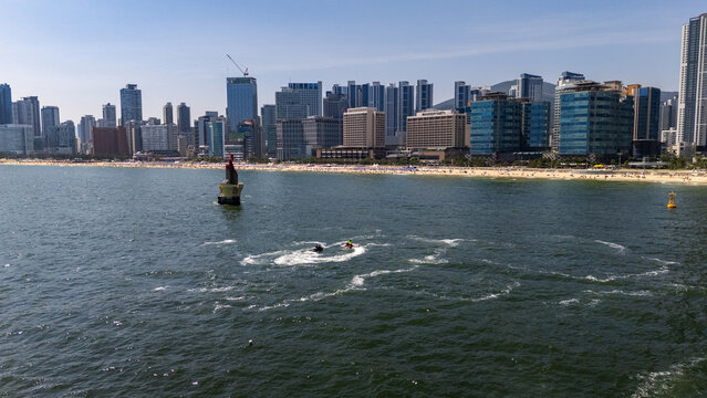Busan, South Korea - 24 June 2026: Aerial view of Haeundae Beach with its sandy shoreline, modern high-rise buildings in the background, and jet skis creating circular wakes in the water.