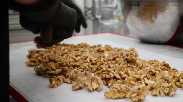 close-up shot of a pastry chef's hands in black gloves using a large kitchen knife to finely chop a heap of fresh, golden walnut kernels. The chef is preparing ingredients on a white silicone baking m
