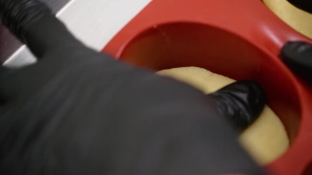 A professional close-up shot capturing a pastry chef wearing black latex gloves carefully shaping and pressing a pale yellow dough or filling into a red, oval-shaped silicone dessert mold. Ideal for c