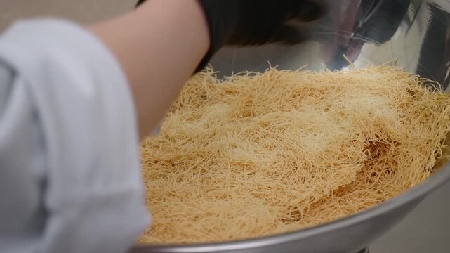 Close-up of a chef's hand in a black glove preparing fine, golden strands of kataifi dough in a large metal bowl. This traditional shredded phyllo pastry is the base for popular Mediterranean and Arab