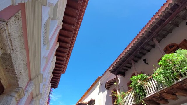 A low angle view looking up at historic Spanish colonial buildings with wooden balconies and lush green plants against a clear blue sky in Walled City Cartagena Colombia
