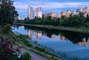 Fototapeta premium Cityscape revealing a calm river reflecting modern and traditional buildings, with lush green trees lining the banks and people enjoying fishing along the water's edge