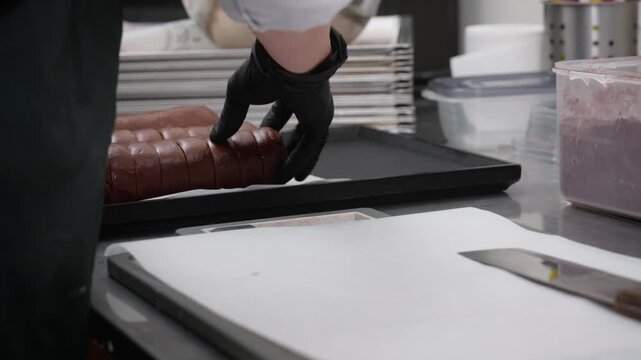 close-up shot of a professional confectioner or pastry chef wearing black nitrile gloves, carefully handling and placing a long, brown cylindrical dessert pastry roll onto a black baking tray. The sce