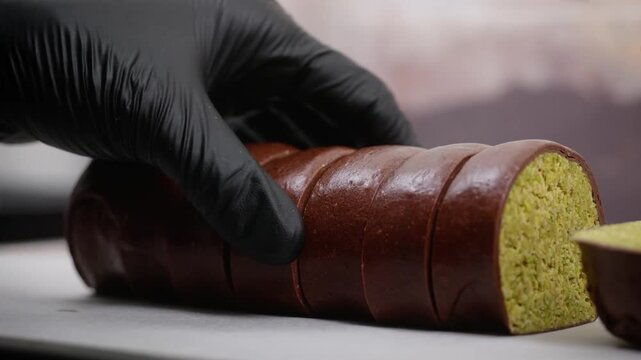 A close-up shot of a pastry chef in black gloves slicing a luxurious chocolate-coated dessert log to reveal a vibrant, crunchy pistachio and kataifi pastry filling.