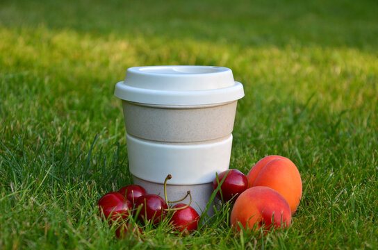 White-beije reusable coffee cup with lid placed on green grass with fresh cherries and peaches. Resting, summer picnic in the garden lawn concept . 