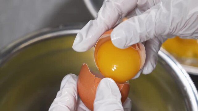 High-angle close-up of a person wearing white gloves carefully separating a raw egg yolk from the white into a metal bowl, essential for making professional custards, pies, and pastries.