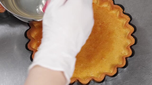 Close-up of a chef wearing white gloves using a pink pastry brush to apply a glossy glaze to a perfectly baked, golden-brown shortcrust tart shell in a fluted tin.