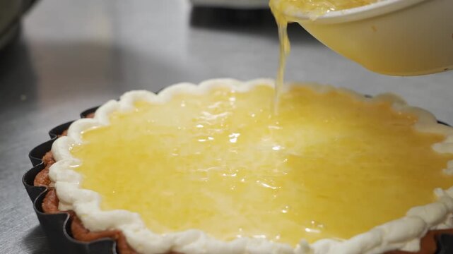 High-angle close-up footage of a professional chef pouring a glossy, yellow lemon curd filling from a white bowl onto a smooth cream-layered pie in a fluted tart pan. The scene highlights the rich tex