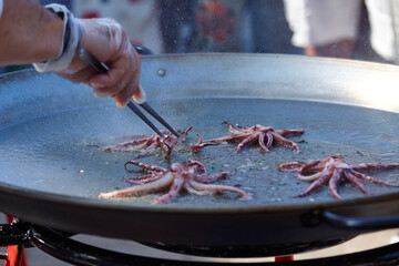 Seafood paella preparation at an open-air festival with a chef using tongs to handle octopus in a large paella pan over a flame © Izi18