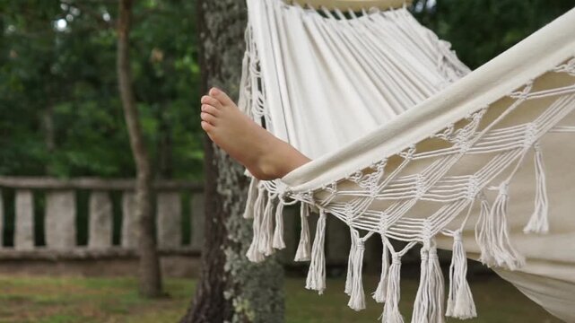 Close-up view of a person's foot resting on the edge of a white fringe hammock as it sways gently against a blurred green park background.