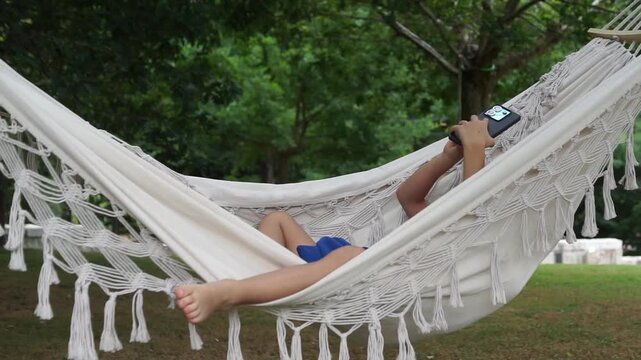 Wide shot of a young boy lying down comfortably in a white macrame hammock swinging in a green park, deeply focused on his smartphone screen.