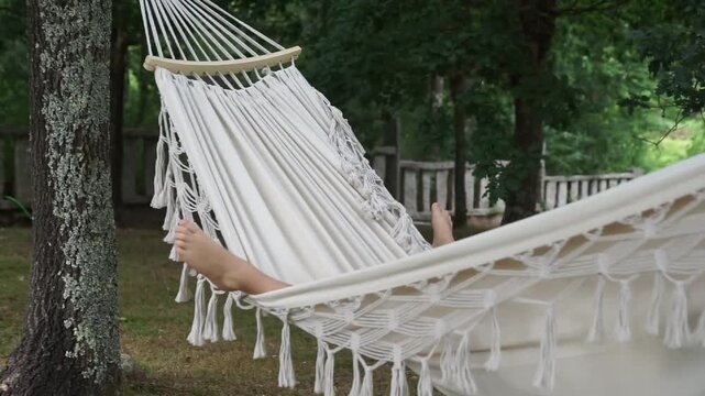 Medium shot of a beautiful white macrame hammock swinging gently in the wind under tree shade, with a person's feet visible inside.