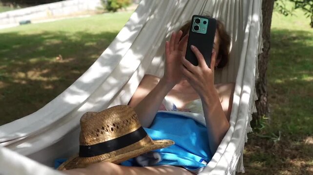Medium shot of a woman lying down comfortably in a white tassel hammock under green trees, browsing social media on her smartphone during vacation.