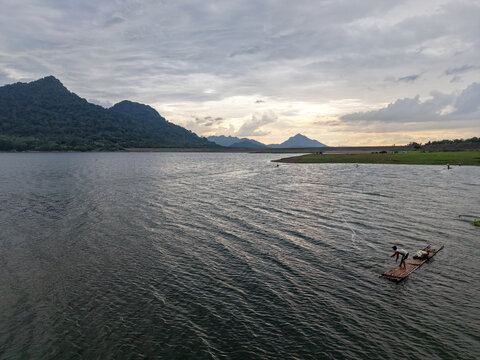 Aerial view of Parang Gombong at Jatiluhur Reservoir with a fisherman on a bamboo raft, calm water, distant mountains, and cloudy sky in Sukasari, West Java, Indonesia.