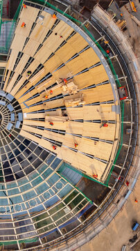 Aerial view of a circular construction site featuring wooden roof panels and metal framework with workers in high-visibility vests working on the structure in Milan, Lombardy, Italy.