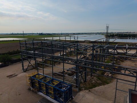 Aerial view of a steel frame structure under construction, with a blue and yellow modular unit on the ground, surrounded by agricultural fields and water in Milan, Lombardy, Italy.