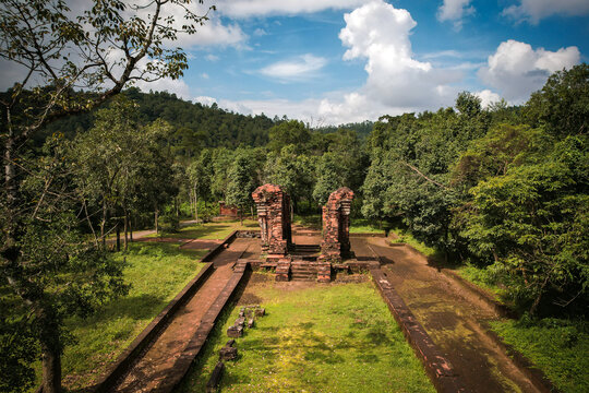 Aerial view of My Son Sanctuary featuring ancient red brick ruins surrounded by lush green forest and trees under a blue sky in Thu Bon, Da Nang, Vietnam.