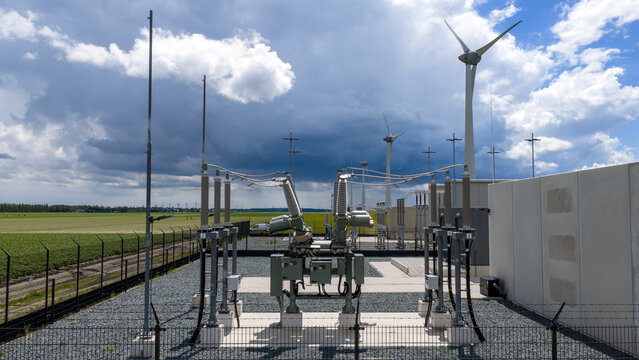 Aerial view of an electrical substation with high-voltage equipment and wind turbines in the background, set amidst green agricultural fields in Espel, Flevoland, Netherlands.
