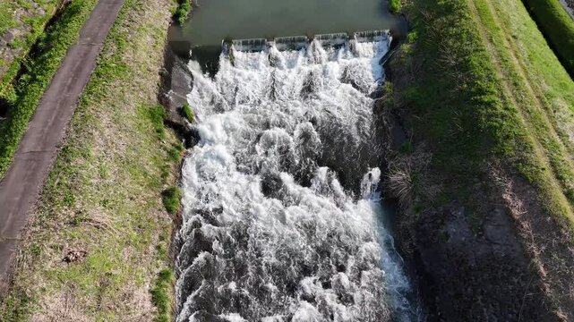 Aerial view of water rushing over a small dam. Green banks frame the turbulent flow. Road runs parallel to the rushing stream. Natural energy moves powerfully through the channel