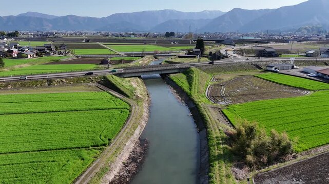 Clear river flows through lush green fields. Mountains stretch far behind the calm waterway. Villages dot the horizon in soft earth tones. Fields glow vibrant under bright, clear skies
