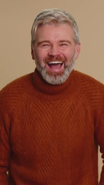 Amused middle-aged man pointing finger to camera, laughing out loud, taunting making fun of ridiculous appearance, funny joke anecdote. Senior mature guy isolated alone on beige studio background