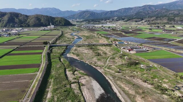Aerial view of rural valley with winding river. Green fields stretch beside the stream. Mountains frame distant horizon. Bridge crosses water calmly. Sunlight bathes landscape in soft glow