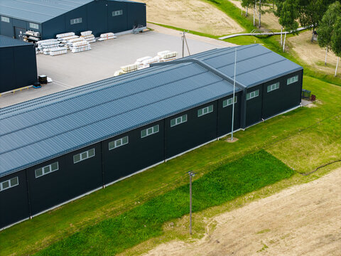 Aerial view of a modern industrial warehouse with dark metal cladding and a pitched roof, situated in a green landscape in Malpils, Sigulda Municipality, Latvia.
