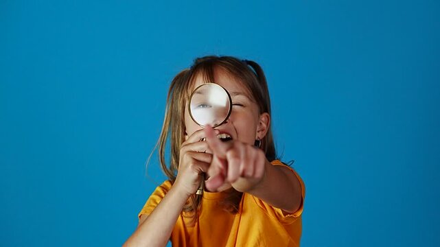 Cute little surprised girl looking through magnifying glass and showing thumbs up against blue background