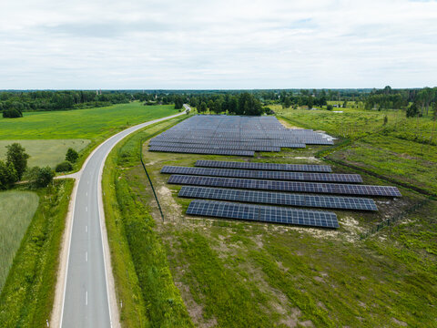 Aerial view of a solar farm with rows of photovoltaic panels situated next to a rural road surrounded by green fields and trees in Suntazi, Ogre Municipality, Latvia.