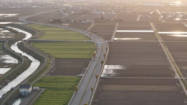 Aerial view of rural countryside at golden hour. Fields stretch in neat rows beside winding canal. Road curves gently through green and brown earth. Sunlight glows softly over distant horizon