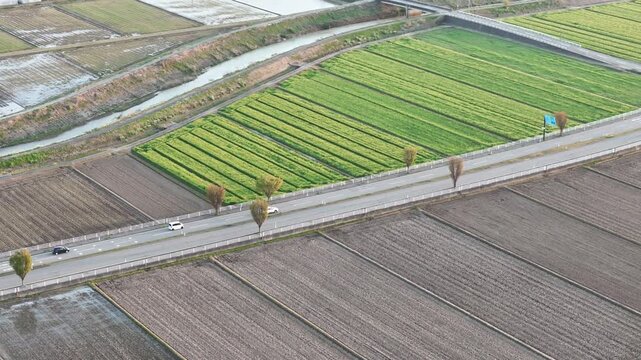Aerial view shows rural landscape with winding road. Green crops contrast with brown fields along the path. Road curves gently through cultivated land with sparse trees. Vehicles move slowly