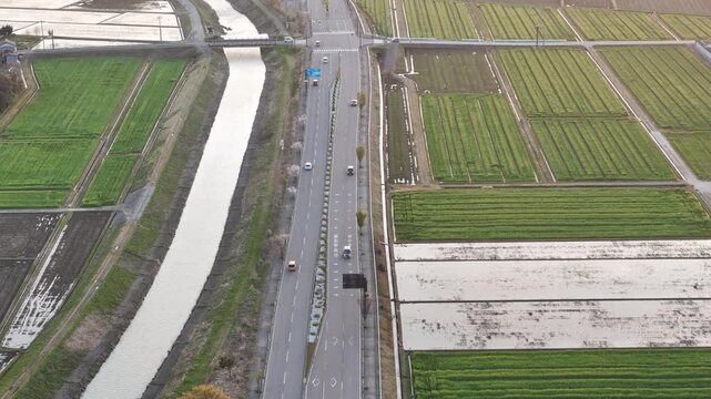 Aerial view of rural road crossing a canal. Green fields stretch wide on either side. Vehicles move slowly on the narrow highway. No people visible in the scene