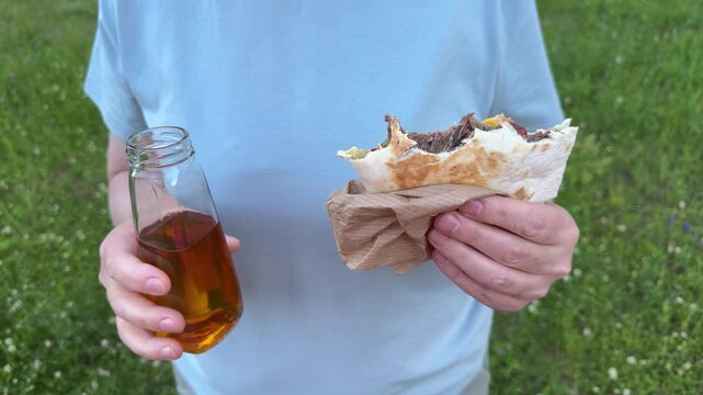 Hands holding a grilled meat wrap and a glass bottle of apple juice, a close-up of food and drink for an outdoor snack