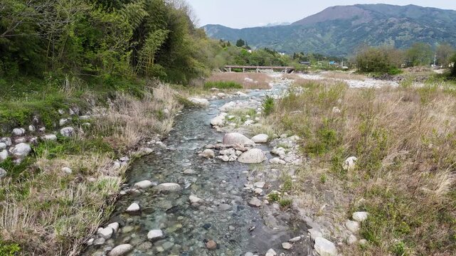 Clear stream flows over stones in quiet countryside. Green grass and trees line the water's edges. Mountains rise gently in the distance. No people or animals appear in the scene
