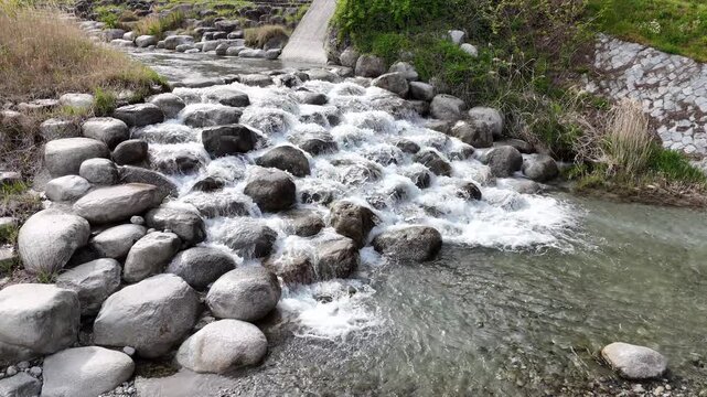 Clear water rushes over smooth, gray stones. Whispering current flows through rocky streambed. Green grass lines banks on either side. No people, no animals, no signs of human activity