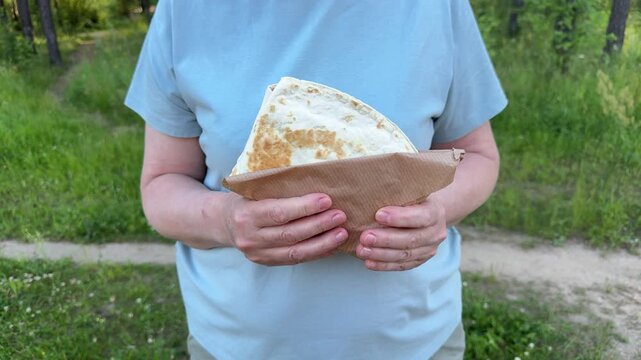 A woman holds a grilled tortilla wrap wrapped in brown paper outdoors, a close-up of a picnic snack in the park and street food