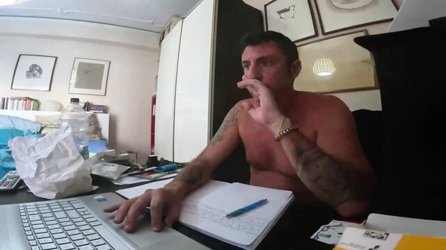 Slow-motion of a man studying in front of a computer and a notebook while sitting at a desk. The man is forty-two years old and bare-chested