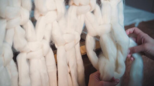 Close-up of a young woman's hands tying macrame knots with wool roving for a decorative tapestry, showcasing handmade textile art, craftsmanship and DIY creativity.
