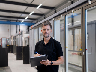 Fototapeta premium Quality worker stands centred writing on a clipboard among glazed door units, a neutral focused look in a production hall.