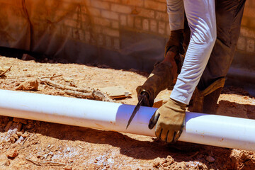 Handyman uses saw to cut white pipe on construction site with dirt, tools around him