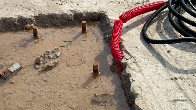 Square excavation next to coiled cable, red conduit end visible on asphalt edge, compact dirt base for bolt anchor, electrical